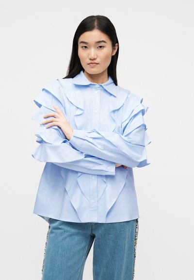 Γυναίκα με μακριά σκουρόχρωμα μαλλιά, φοράει ανοιχτό μπλε μπλούζα με βολάν και μπλε τζιν με στριφώματα στο πλάι διακοσμημένα με πολύτιμους λίθους, με σταυρωμένα τα χέρια, σε λευκό φόντο.