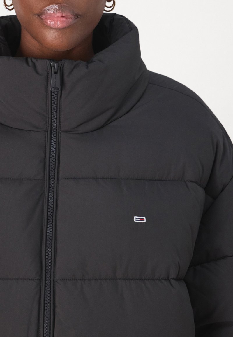 Giacca a vento nera con colletto alto, chiusura a zip e un piccolo logo. Il tessuto è liscio con sezioni trapuntate per l'isolamento.