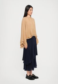 Nainen seisoo sivuttain päällään löysä beige paita, jossa on röyhelöhihoja ja kerroksellinen tummansininen hame, yhdistettynä mustiin loafereihin.