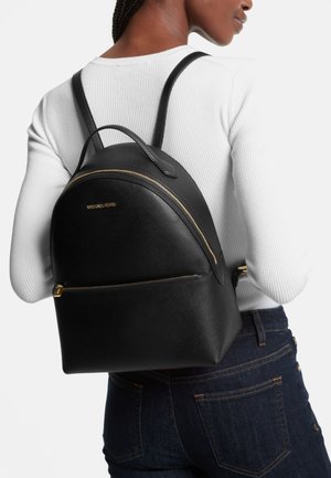 Naine kannab musta Michael Kors nahast seljakotti kuldsete tõmblukkude ja logoga, mida kannab valge soonikkudega topp ja tumedate siniste teksadega.