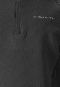 Haut performant noir avec une finition texturée, doté d'une fermeture zippée à mi-hauteur et d'un logo métallique "ENDURANCE" sur la poitrine.