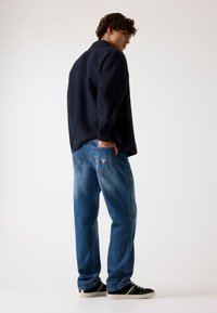 Marineblaues Fleece-Hemd mit Kragen, kombiniert mit hellblauen Jeans mit Gesäßtaschen und schwarzen Turnschuhen mit weißen Sohlen.