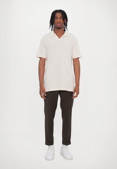 Polo beige à manches courtes avec un col en V et une texture douce, associé à un pantalon fuselé marron foncé et des baskets blanches.