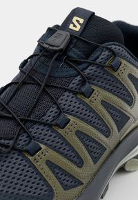Navy and olive mesh athletic shoe featuring a textured upper, elastic laces, and a reinforced heel for support and durability.