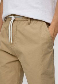 Beige leichte Hose mit Kordelzugbund, Seitentaschen und glatter Textur. Getragen mit einem schlichten weißen T-Shirt.
