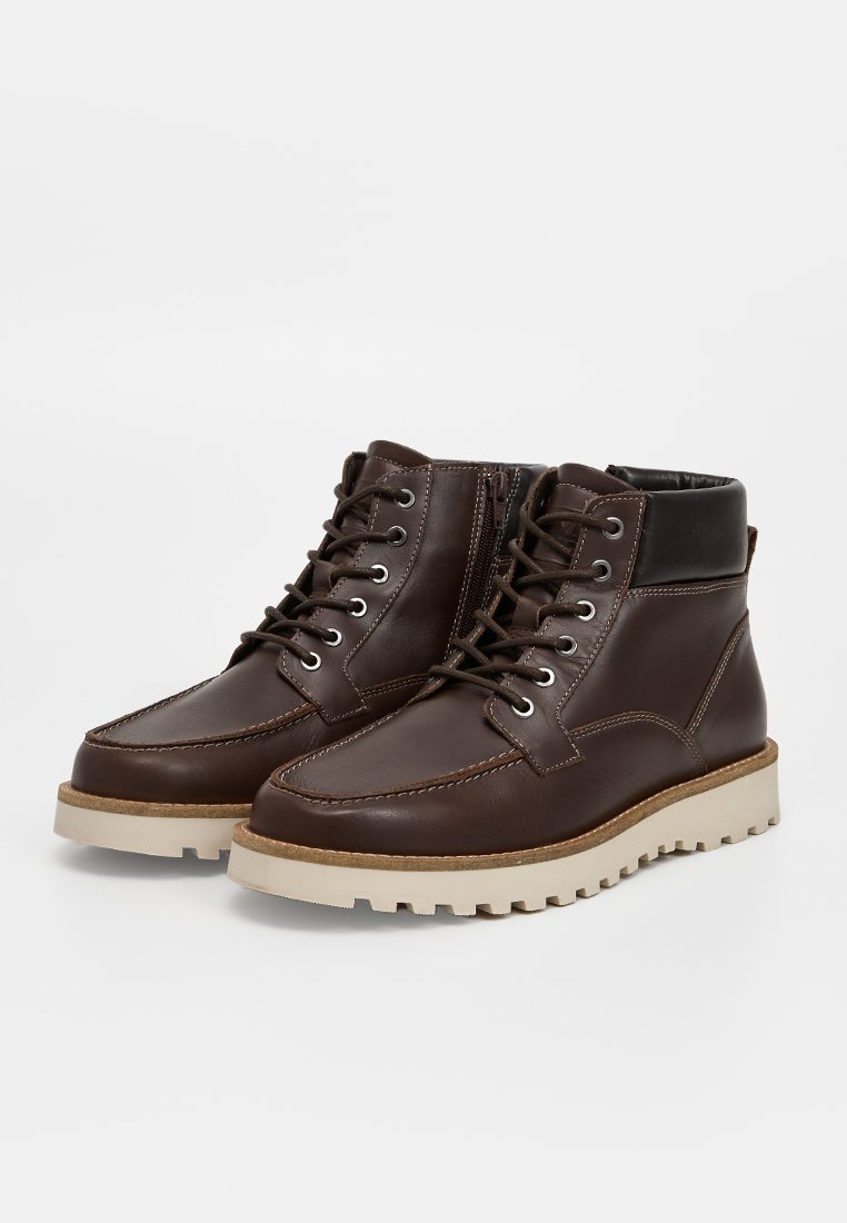 Braune Leder-Mid-Boots mit runder Spitze, kontrastierender, tanfarbener Gummisohle, Metallösen und Schnürung auf der Vorderseite; mit gepolstertem Knöchel.