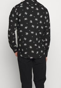 Camicia nera a maniche lunghe con piccolo motivo floreale bianco, design con bottoni e orlo curvo. Il materiale sembra leggero e morbido.