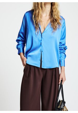 Person wearing bright blue satin button-up shirt with loose brown trousers holding black handbag against white background.