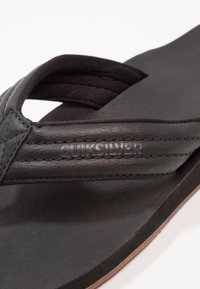 Sort sandal i svart med skinnøvre, teksturerte stropper og polstret såle. Logo vist på siden med preget skrift.