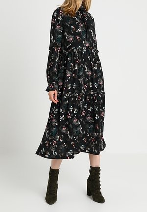 Kvinde iført en sort blomsterprintet langærmet midi-kjole med lagdelt nederdel og olivengrønne snørelåsede hælede ankelstøvler.