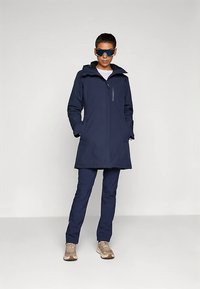 Marineblå vandtæt jakke med lynlås foran, høj krave og sidelommer, kombineret med matchende slim-fit bukser og lyse sneakers.