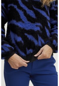 Svetrov s črno-modrim vzorcem in puhasto teksturo ter rebrastim robom, v kombinaciji z oprijetimi modrimi hlačami, z rokami, ki prilagajajo rob.