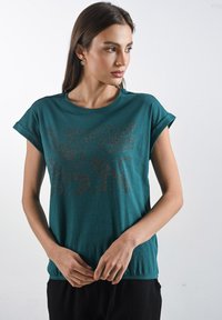 Mladá žena s dlouhými vlasy nosí tyrkysové tričko s dekorativním vzorem. Stojí na světlém pozadí a dívá se do strany.