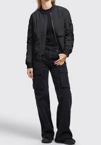 Femme portant une veste bomber noire à fermeture éclair, un shirt noir à manches longues, un pantalon cargo noir avec de grandes poches et des baskets noires sur un fond gris.