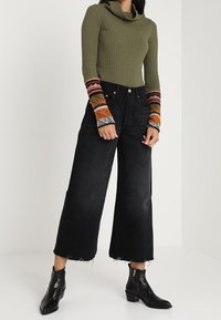 Kvinna i en olivgrön ribbad tröja med flerfärgad mönstrad manschett, svarta vida cropped jeans och svarta ankelstövlar.