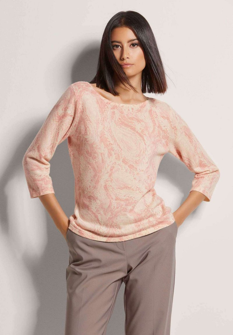 Helles rosa Langarmoberteil mit weicher Textur und abstraktem Wirbelmuster, kombiniert mit grauen, taillierten Hosen. Gestylt vor einem neutralen Hintergrund.