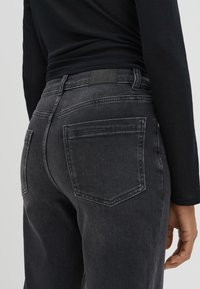 Person wearing black faded jeans and a long-sleeve black top, shown from the back waist to mid-thigh with hand relaxed by side.