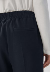 Svart byxor med elastisk midja och en enda bakficka, bärs med en vit långärmad topp.