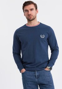 Marineblaues Langarmhemd mit einem rot-weiß gestreiften Kragen, das ein kleines Logo auf der Brust trägt. Weiches Material und klassischer Schnitt.