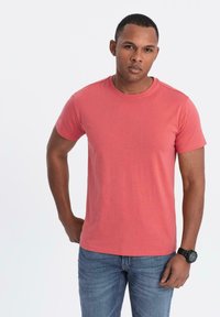 Korallfärgad kortärmad T-shirt i bomull med rund halsringning. Bärs med blå jeans och en svart armbandsklocka. Slät textur.