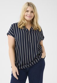 Blouse bleu marine à manches courtes avec des rayures verticales beiges, un col en V boutonné et une coupe décontractée, associée à un jean foncé.