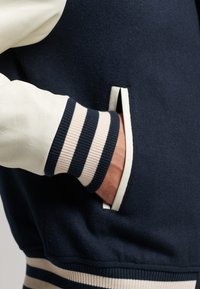 Marinblå ullblandning jacka med krämfärgade läderärmar. Har randiga marinblå och beige ribbade muddar samt sidofickor. Mjuk textur, modern design.