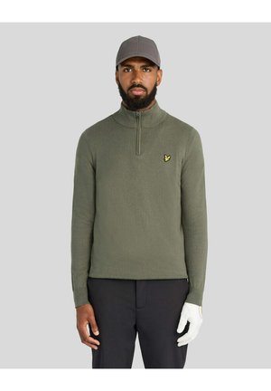 Mann trägt olivgrünen Pullover mit Viertelreißverschluss, graue Kappe, schwarze Hose und einen weißen Golfhandschuh an der rechten Hand, steht vor einem schlichten Hintergrund.
