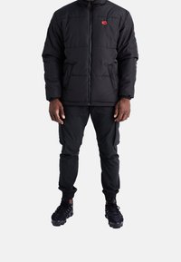 Mann in einer schwarzen gepolsterten Jacke mit einem roten Rosenemblem, schwarzen Cargo-Hosen und schwarzen Sneakers, der vor einem weißen Hintergrund steht.