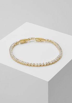 Bracelet de tennis en or avec des pierres rondes claires uniformément disposées le long de sa longueur, présenté sur un socle blanc sur fond gris.