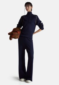 Granatowy dres dziany, składający się z golfu z dekoltem w serek, ściągaczami na rękawach oraz spodni o szerokich nogawkach. Modelka trzyma brązowo-czerwoną, paskowaną torbę.