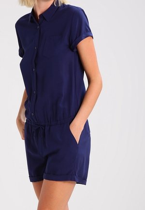 Femme portant une combinaison-short bleu marine à manches courtes avec col, boutons sur le devant, taille à cordon de serrage et revers sur le bas du short, mains dans les poches.