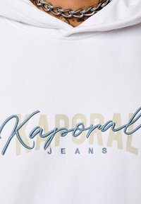 Haut à capuche blanc avec "Kaporal" en broderie bleue et "JEANS" en texte beige clair en dessous. Tissu doux avec une coupe décontractée et capuche.