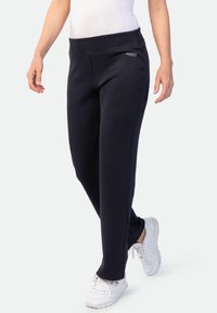 Frau in schwarzen Sporthosen und weißen Sneakers, die mit entspannten Armen an den Seiten nach vorne geht, vor einem einfarbigen Hintergrund.