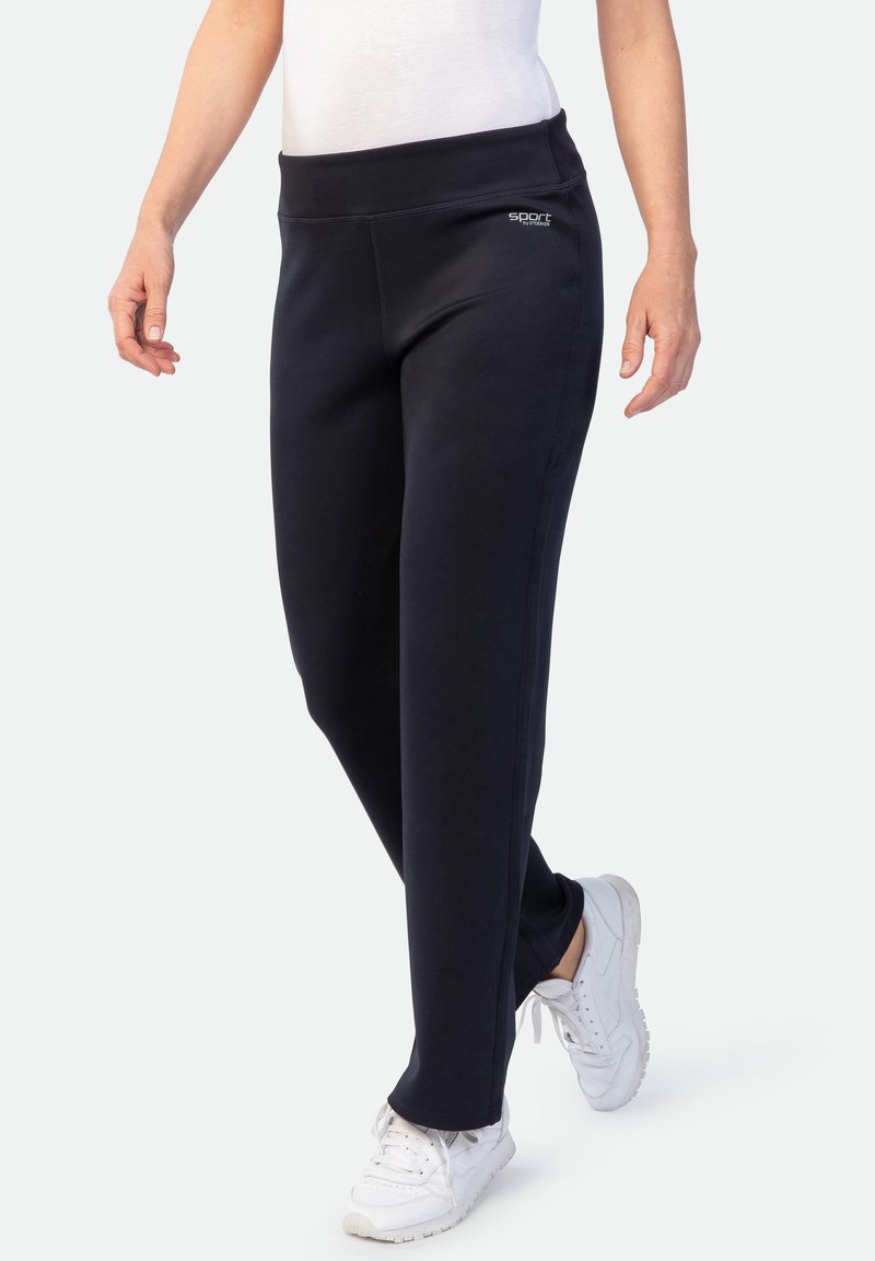 Frau in schwarzen Sporthosen und weißen Sneakers, die mit entspannten Armen an den Seiten nach vorne geht, vor einem einfarbigen Hintergrund.