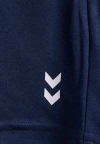 Marinblått sporttyg med en texturerad yta, med en vit triangel-logotyp på vänster sida. Ren sömnadsdetalj vid fållen.