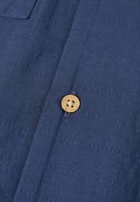 Tissu bleu marine avec des rayures texturées, comprenant un bouton circulaire en bois de couleur fauve à quatre trous fixé sur la patte de boutonnage.