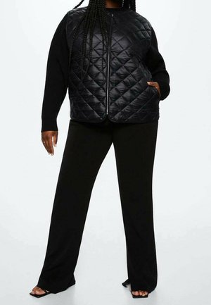 Personne portant un gilet matelassé noir zippé par-dessus un haut à manches longues, un pantalon noir large et des talons ouverts, debout avec une main dans la poche.