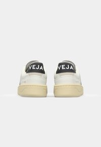Sneaker in pelle bianca con accenti grigi e suole in gomma beige. I talloni neri presentano il logo "VEJA" in bianco. Design pulito, stile low-top.