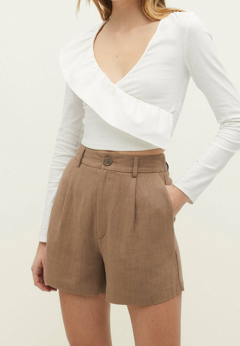 Haut blanc à manches longues avec un design froncé et diagonal, associé à un short en lin marron taille haute, doté d'une fermeture à bouton et de plis.