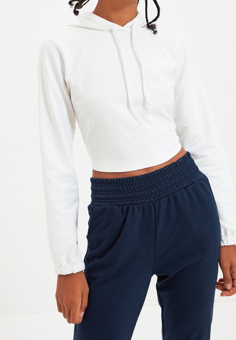 Personne portant un sweat à capuche court blanc et un pantalon bleu marine taille haute avec une ceinture élastique, debout devant un fond blanc.