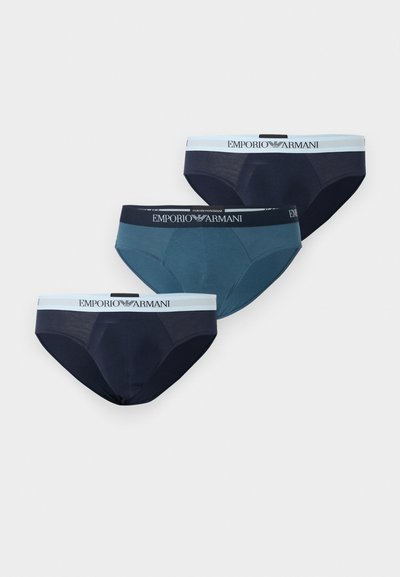 Trois slips pour hommes Emporio Armani en marine et bleu, chacun avec une ceinture élastique à logo, disposés côte à côte sur un fond blanc.