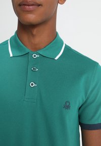 Tyrkysové polo tričko s texturovanou tkaninou, tříknoflíkovou légou, kontrastním bílým límcem a tmavě modrými akcenty na rukávech. Malé logo na hrudi.