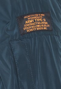 Blouson de vol en nylon bleu marine avec une étiquette noire portant un texte orange, indiquant des détails tels que "INTERMEDIATE ARMY TYPE-U" et des informations sur le fabricant.