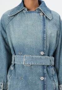 Lyseblå denim trenchcoat med krage, frontknapper og belted midje. Har en teksturert finish og synlige sømdetaljer.