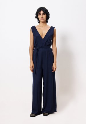 Combinaison marine avec un décolleté en V plongeant, sans manches, une taille ceinturée et des jambes larges. Le tissu a une texture lisse, associé à des chaussures à motifs.