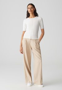 Witte geribde top met gekartelde randen, gecombineerd met lichtbeige wijde broek. Model staat met handen in de zakken, draagt witte sneakers.