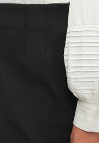 Close-up de calças pretas sob medida e blusa de mangas longas brancas com pregas horizontais nos punhos, vestida no braço de uma pessoa.