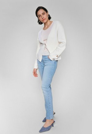 Naine seisab halli tausta ees, kandes valget kardigani, heleroosat topi, helesiniseid teksaseid ja siniseid teravate ninadega madala kontsaga kingi.