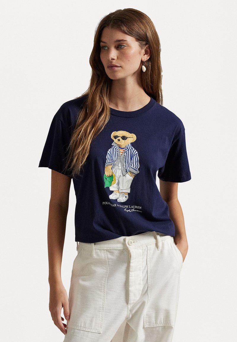 Navy cotton t-shirt featuring a bear graphic in sunglasses, striped jacket, and white pants. Accented with "POLO RALPH LAUREN" text.