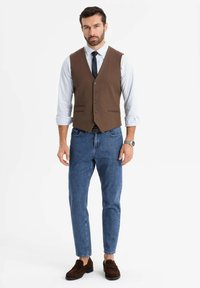 Mann in braunem Weste über einem weiß gestreiften Hemd mit hochgekrempelten Ärmeln, blauen Jeans, dunklen Slippern und einer Armbanduhr vor einfarbigem Hintergrund.